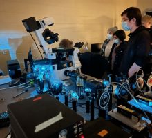 Several scientists in a darkened room that contains a laser table and a microscope.