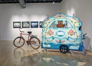 A fanciful 2-wheeled trailer pulled by a bicycle on display in an art museum