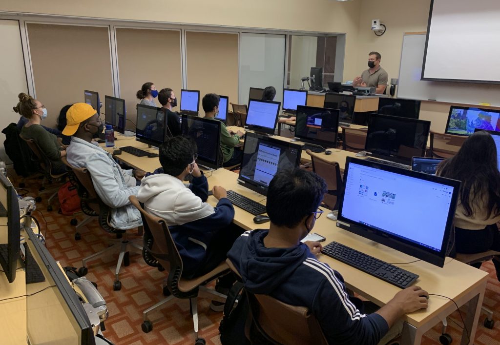 An instructor gesticulates to a computer lab full of students seated at computers.
