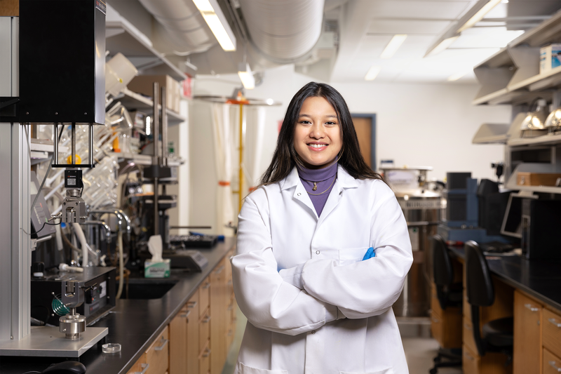 A smiling researcher stands in a lab, wearing a lab coat.