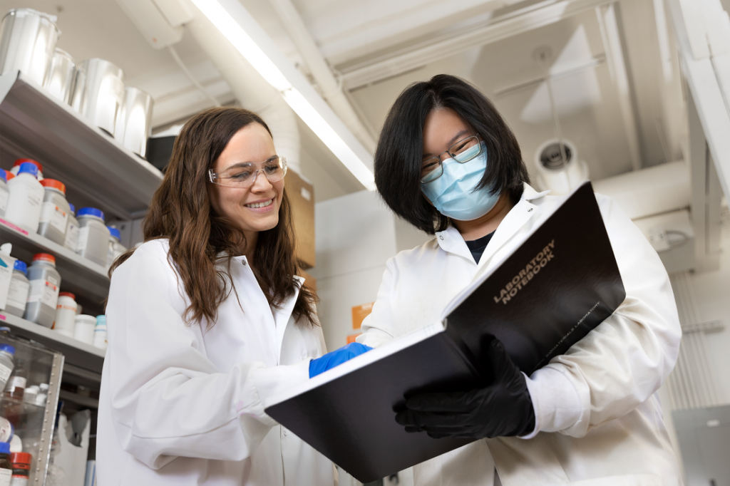 Professor Monroe examines a page in a lab notebook held by a student in the laboratory