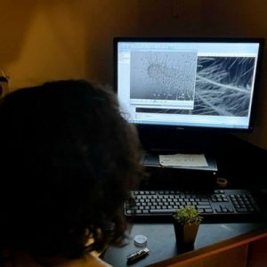 A person sits in front of a computer monitor in a darkened room using a scanning electron microscope