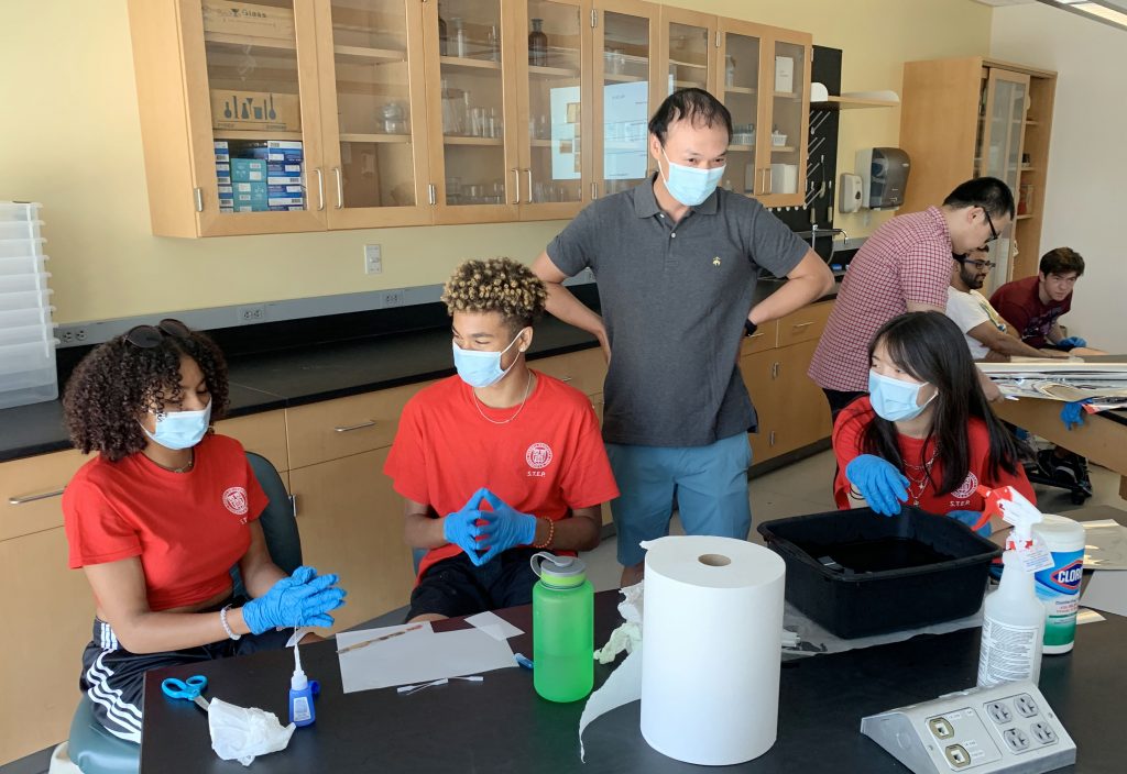 A professor oversees a hands-on activity with several high school students in a lab