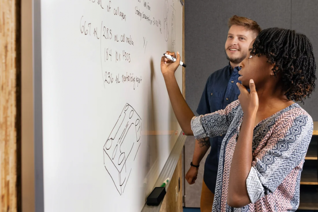 Two students draw on a white board in a conference room.