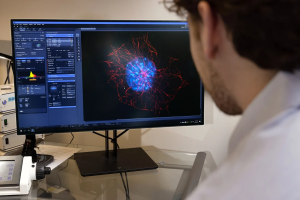 A student examines a computer monitor showing multicolored microscopy of a cell.